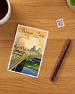 Postcard with cityscape design on a wooden surface with a pen and stamp.